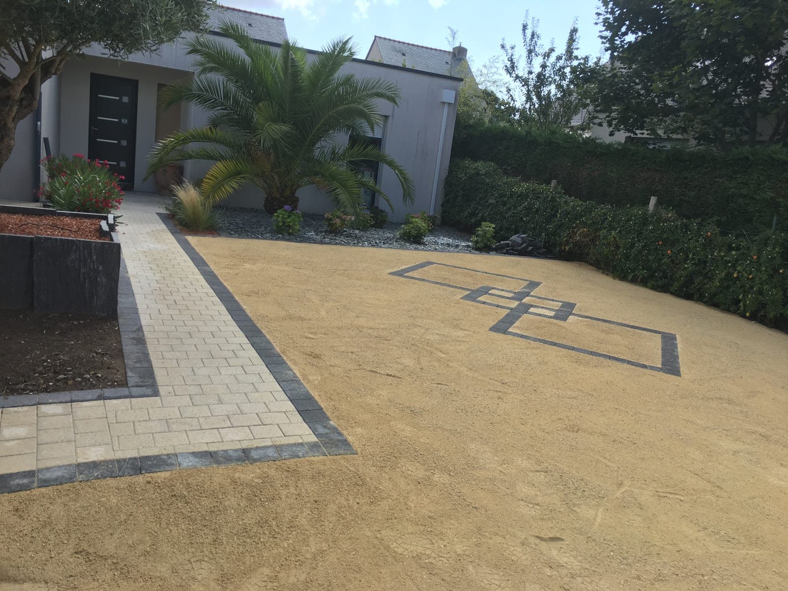 Cour en revêtement stabilisé couleur sable ornée d'un motif de pavés anthracite, à côté d'une allée en pavés clairs